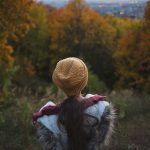 girl in parka and yellow hat stands in Park with her back to camera